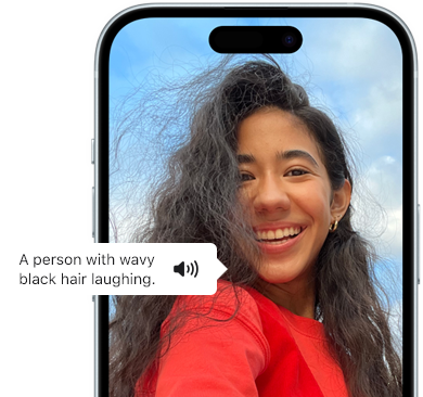 iPhone 15 se zobrazeným oznámením funkce VoiceOver, která popisuje fotografii: smějící se osoba s vlnitými černými vlasy